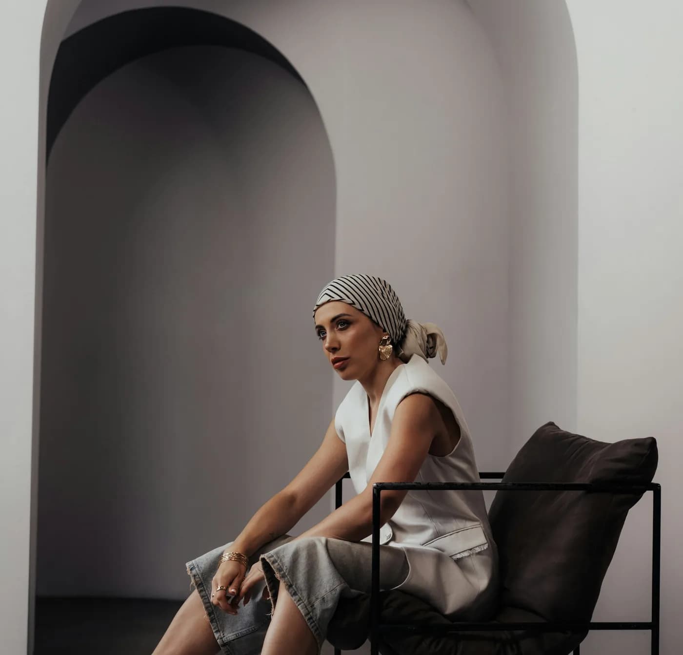A woman seated in a low chair inside a plaster arch, in a cream vest and wide trousers