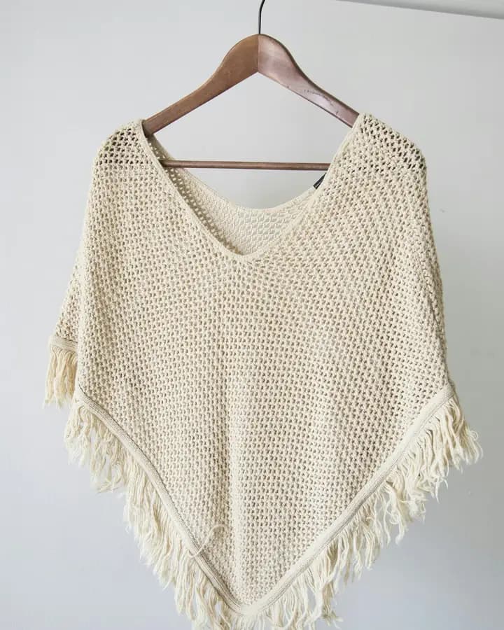 Cream knitted poncho with a fringed hem, on a wooden hanger