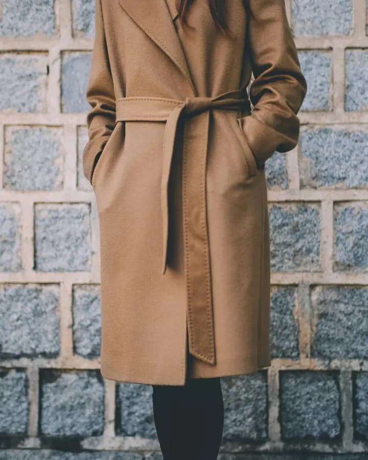 A camel wrap coat worn open, photographed against a stone wall