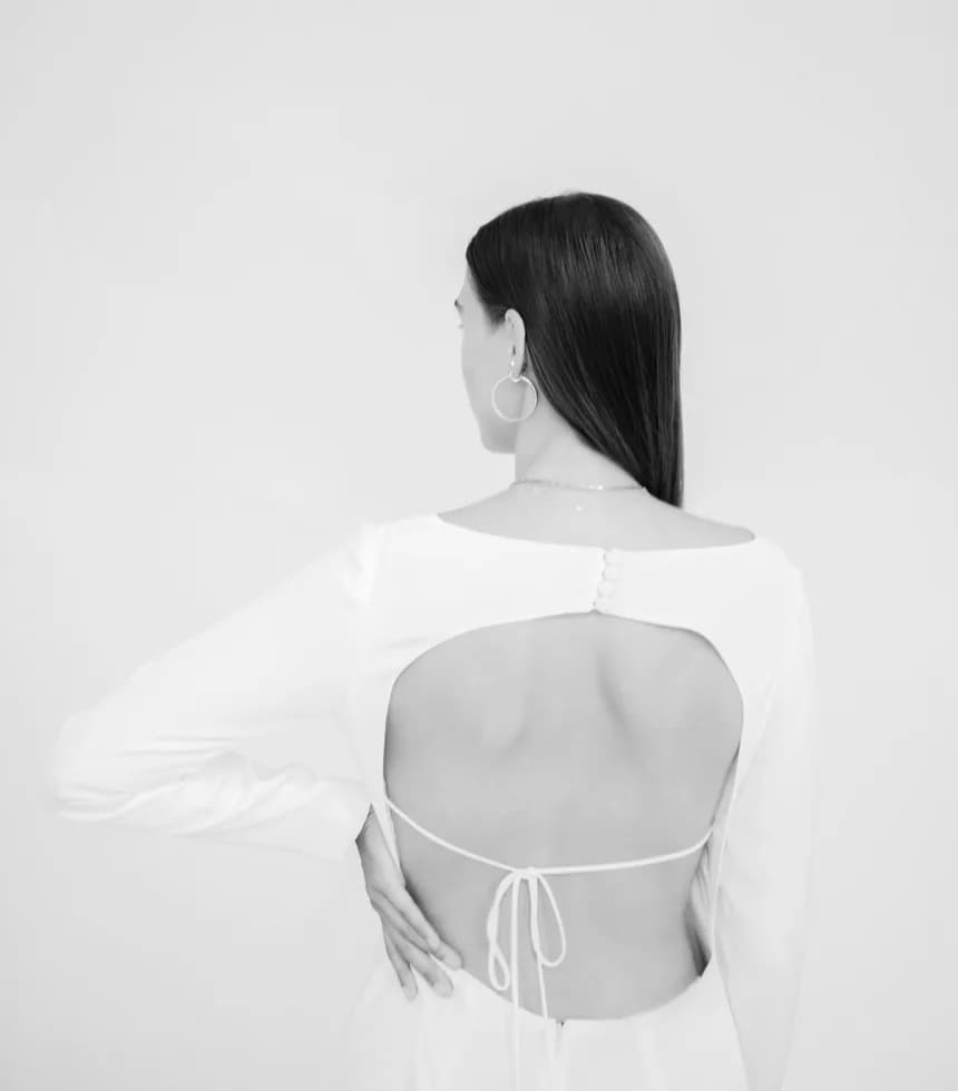 A woman photographed from behind in a white open-backed top, against a pale wall