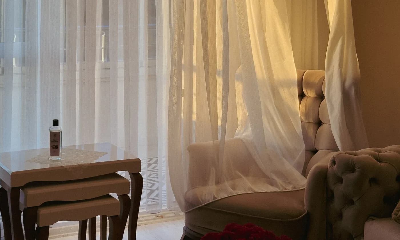 Sunlight through sheer linen curtains onto a pair of upholstered chairs and a nest of side tables